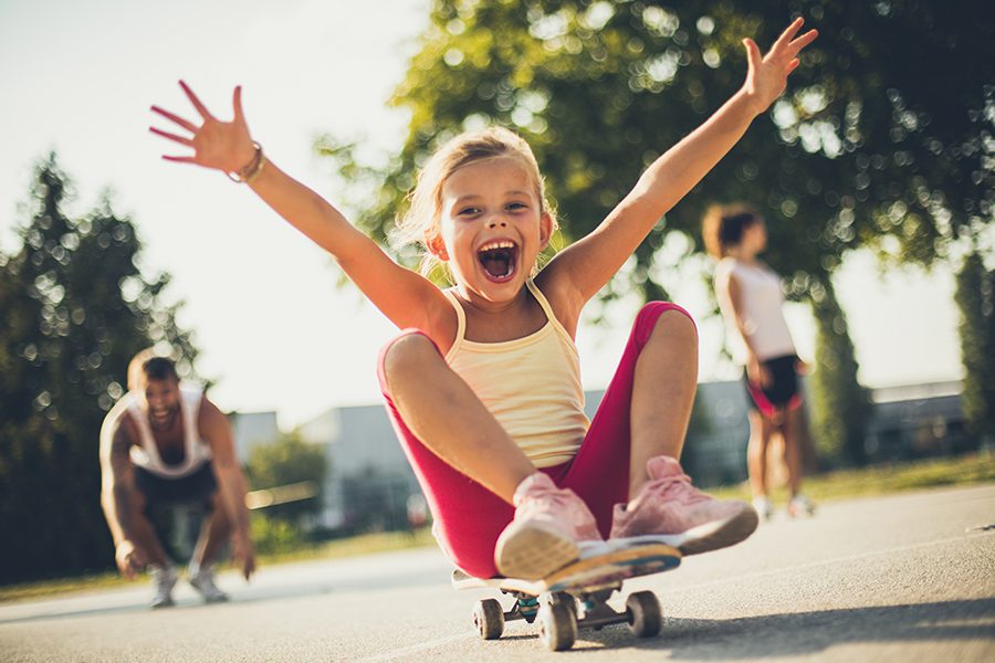 Employee Benefits - Little Girl Having Fun and Sitting on a Skateboard with Parents Blurred in the Distance on a Sunny Day